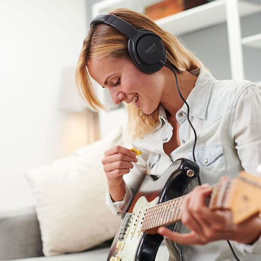 Picture of woman wearing RH-5 Roland Headphones while playing an electric guitar and sitting on a couch.